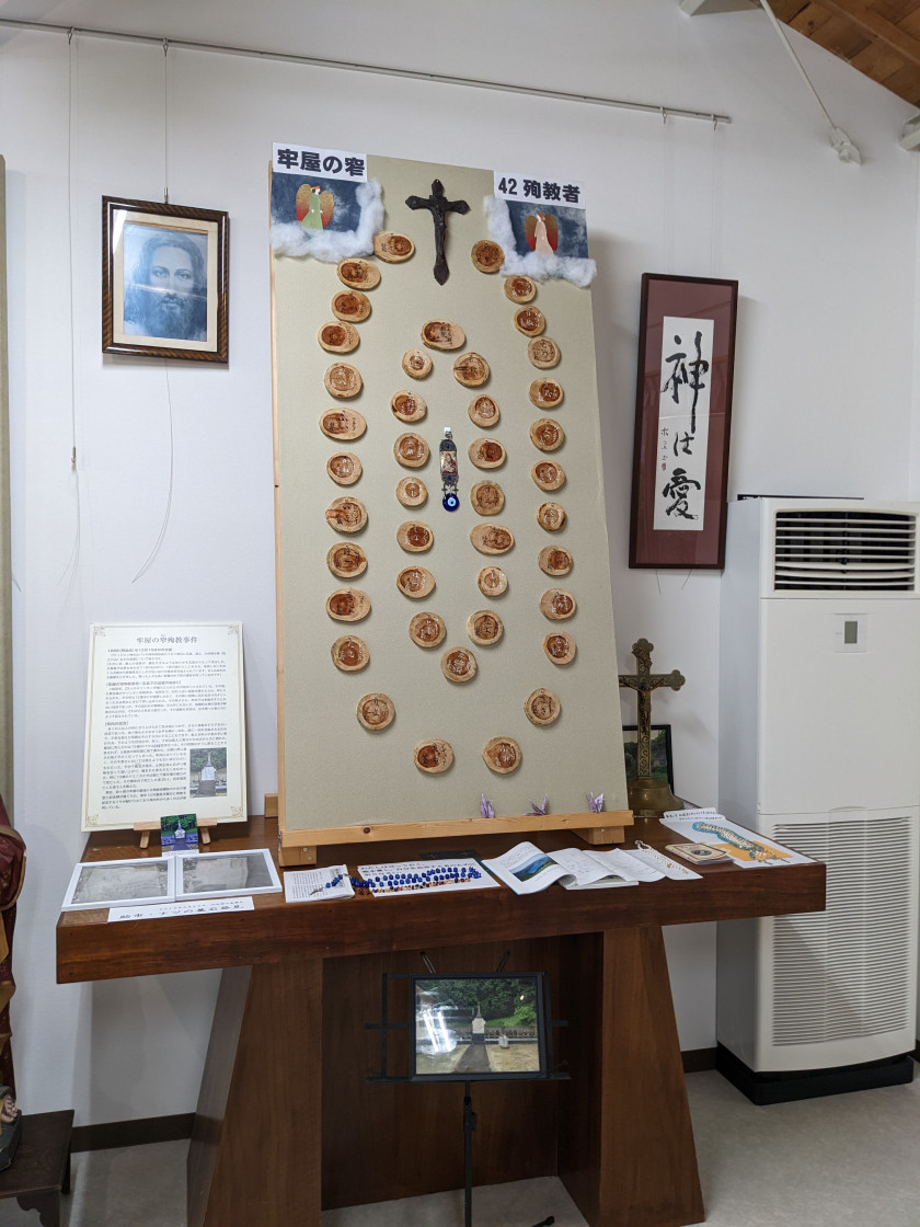 A memorial exhibition of Roya no Sako at the Catholic Museum on Hisaka Island. A thick wooden table is topped with papers and books along with a tall board featuring several dozen small circular plaques and a cross.