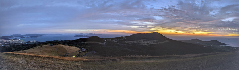 A photograph showing a wide shot of an open, hilly landscape at sunset. It is the view from Onidake on Fukue Island.