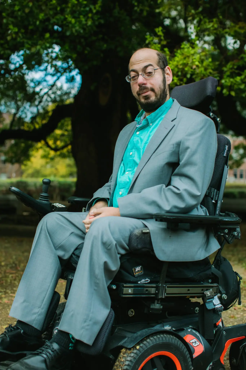 Professional headshot of the late Mark Bookman, in bright blue-green shirt and pale grey suit, sitting in a motorized wheelchair.