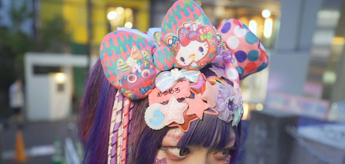 Top part of a woman's head with dyed purple hair, and many hair accessories, including star-shaped hairclips in pink and white, and a large plush bow decorated with the character Hello Kitty