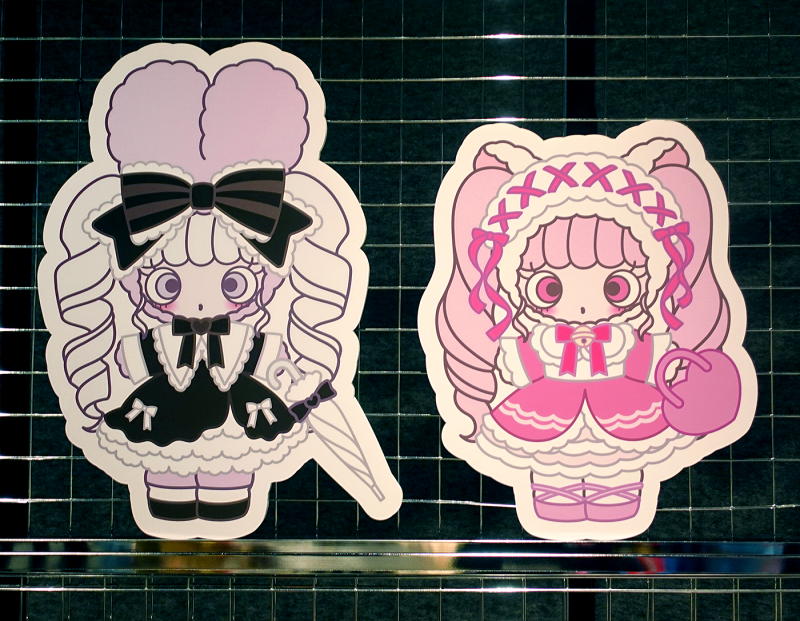 Two stickers of cute female characters on a black gridded background in frilly outfits with bows, the character on the right is dressed in pink and the character on the left in black