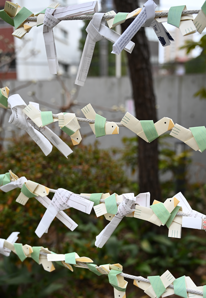 Paper strips folded and tied to cords in four horizontal rows across the image, some papers are yellow and have been decorated to resemble birds, vegetation in the background