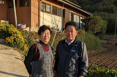 Miyamoto Fujie and Jitsuo stand in front of their home in Hamawaki, Hisaka Island. Middle-aged Japanese couple in front of a brick home.