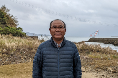 A photograph of a partial view of Fr. Nakamura Mitsuru, Fukue Island, A middle-aged Japanese man with glasses in dark blue puffer jacket stands in front of a grassy area in front of a body of water.
