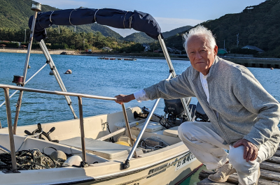 A photograph of Kakimori Kazutoshi with boat on Naru Island. Senior Japanese man in grey jacket kneeling beside a white boat on a sunny day.