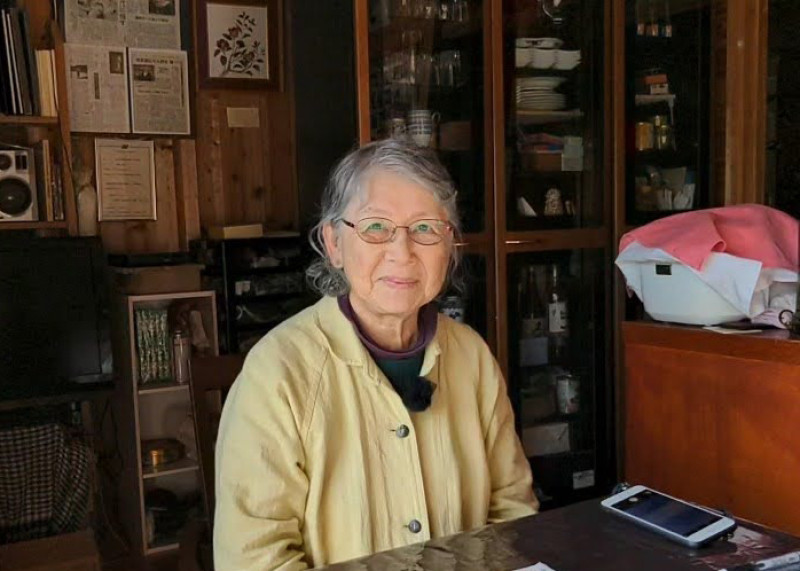 A photograph of Urakami Sachiko in front of shelves at the Hidden Christian Museum, Naru Island. An older Japanese woman with glasses in a pale yellow sweater, smiling.