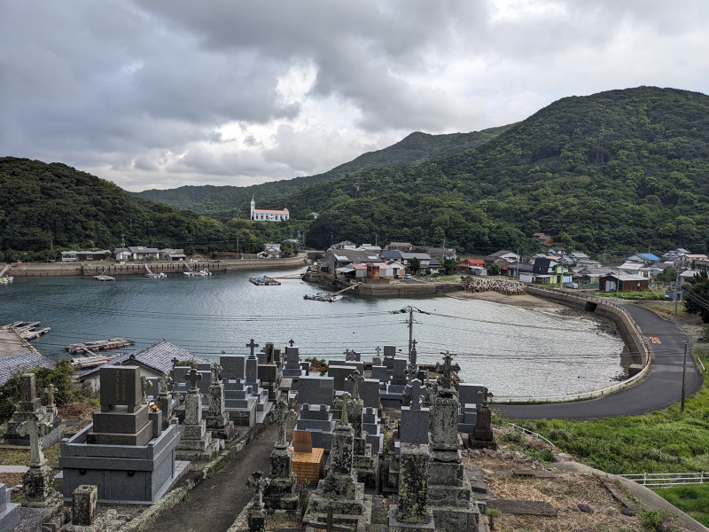 A photograph of of Kiri church and cemetery, Nakadōri Island, Upper Gotō. In the foreground is a series of gravestones, with a circular body of water in the middleground set in front of a red-roofed church sitting on the forested mountainside.