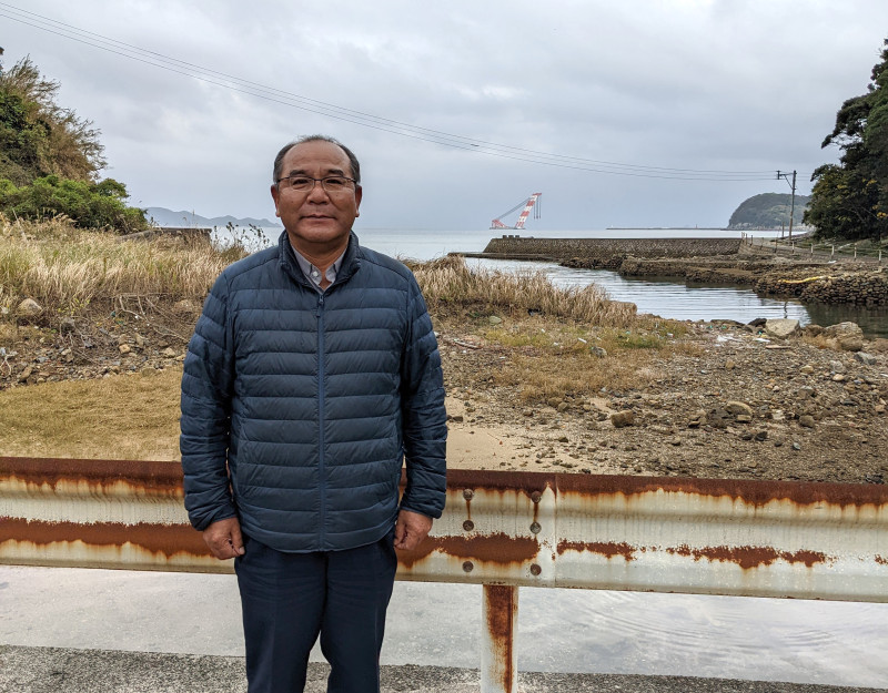 A photograph of Fr. Nakamura Mitsuru, Fukue Island, A middle-aged Japanese man with glasses in dark blue puffer jacket stands in front of a grassy area in front of a body of water.