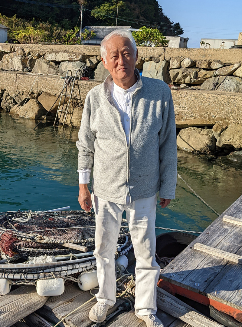 A photograph of Kakimori Kazutoshi on Naru Island. A senior Japanese man in grey jacket stands on a small wooden dock in the water.