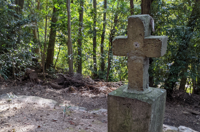A photograph with a partial view of grey gravestone with cross at Shinto shrine (Karematsu Jinja) against green forest background. The grave is carved with the name Maria in katakana.