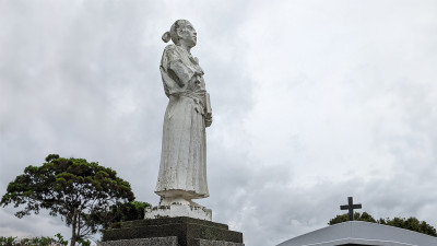A photograph of a partial view of a stone statue of Yohane Gotō found on Fukue Island at Mizu-noura Church. The statue is of a man in kimono holding a cross to his chest and a book at his side, staring into the distance.