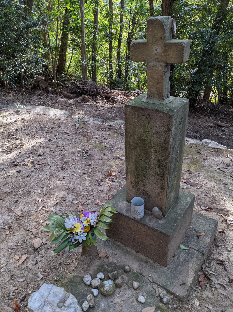 A photograph of a grey gravestone with cross at Shinto shrine (Karematsu Jinja) against green forest background