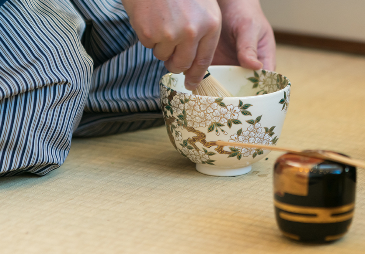 Partial view of a tea practitioner holding a tea bowl and whisk with a tea caddy and ladle sitting on a tatami mat.