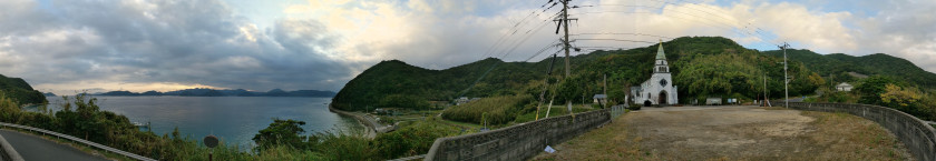 Panoramic photo of the Hamawaki Church and surrounds, a green roofed white colored church against green forest background with a body of water to the left.