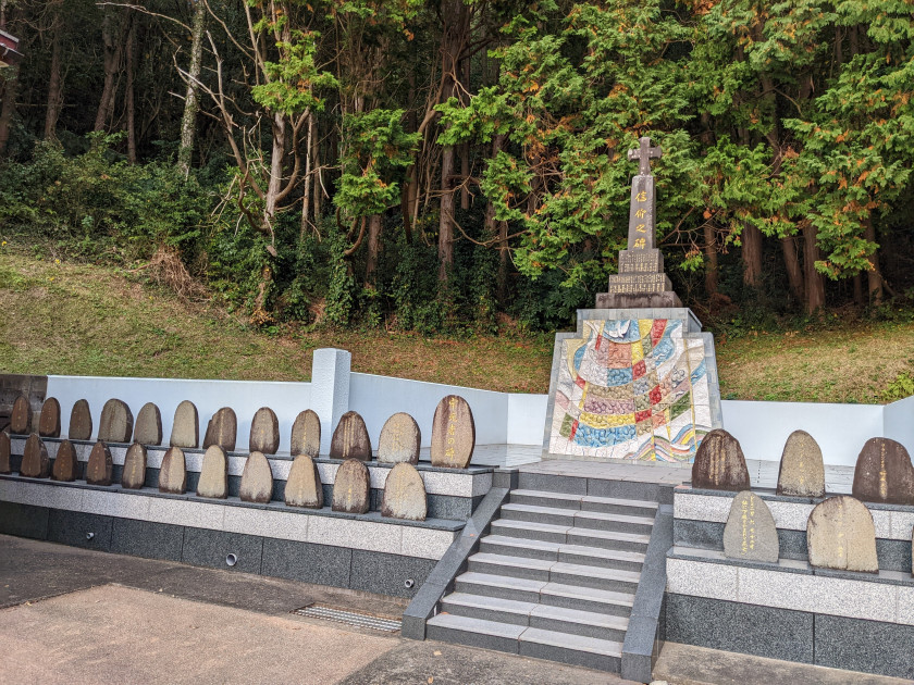 Photograph of a memorial site with two rows of stone plaques in front and a taller statue in the back. The thicker base is a colorful mosaic and the top is a rectangular stone with gold writing topped with a stone cross.