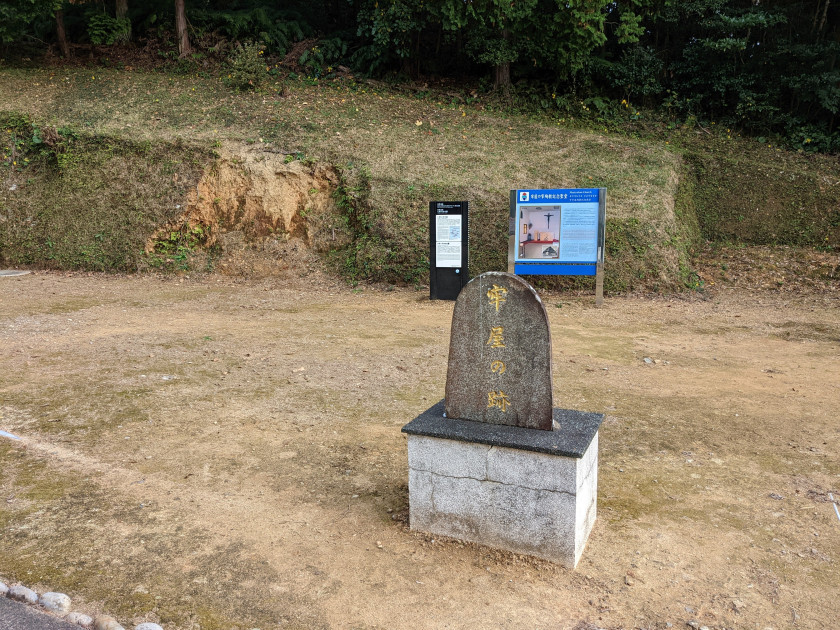 A photograph of the Rōya no sako site on Hisaka Island. A stone plaque with 牢屋の跡 written in gold, sitting in empty field.