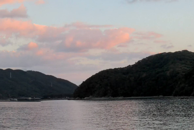 A photograph of the view from Hidden Kirishitan Resource Center, a sunset over two small mountains with a placid body of water in front.