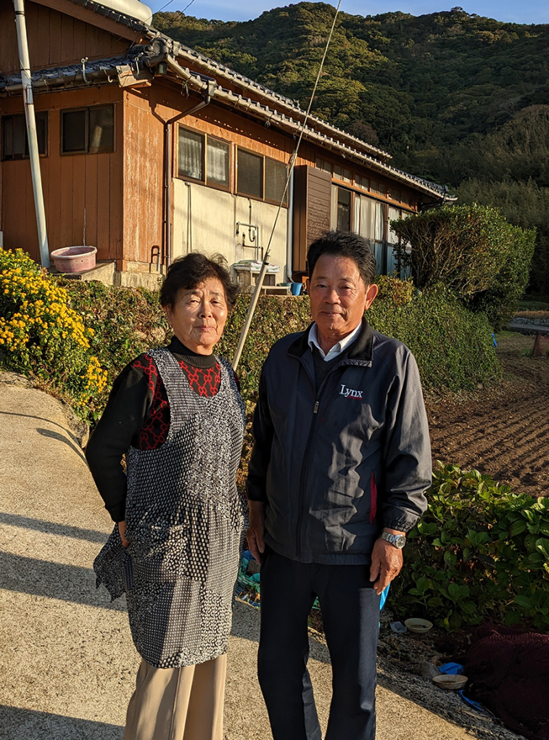 A photograph of Miyamoto Fujie and Jitsuo standing in front of their home in Hamawaki, Hisaka Island. Middle-aged Japanese couple in front of a brick home.