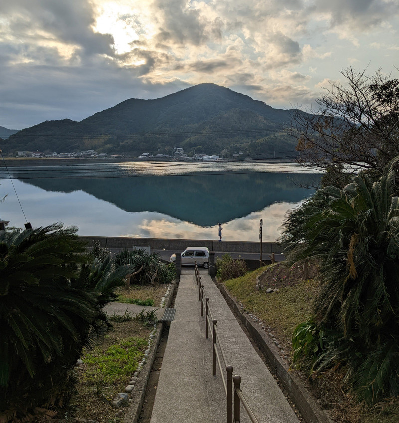 A photograph of the view from the martyrdom site looking toward the old magistrate’s house across the harbour, Rōya no Sako. The clouds of a late afternoon float over a scenic mountain reflected over a body of water.