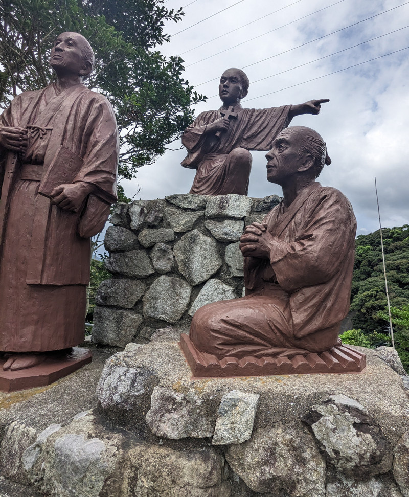 A photograph of a three bronze statues, one kneeling on triangular wedges to represent sangizeme torture. Found outside Kiri Church, Nakadōri Island.