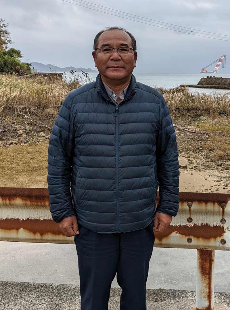 A photograph of Fr. Nakamura Mitsuru, Fukue Island, A middle-aged Japanese man with glasses in dark blue puffer jacket stands in front of a grassy area in front of a body of water.