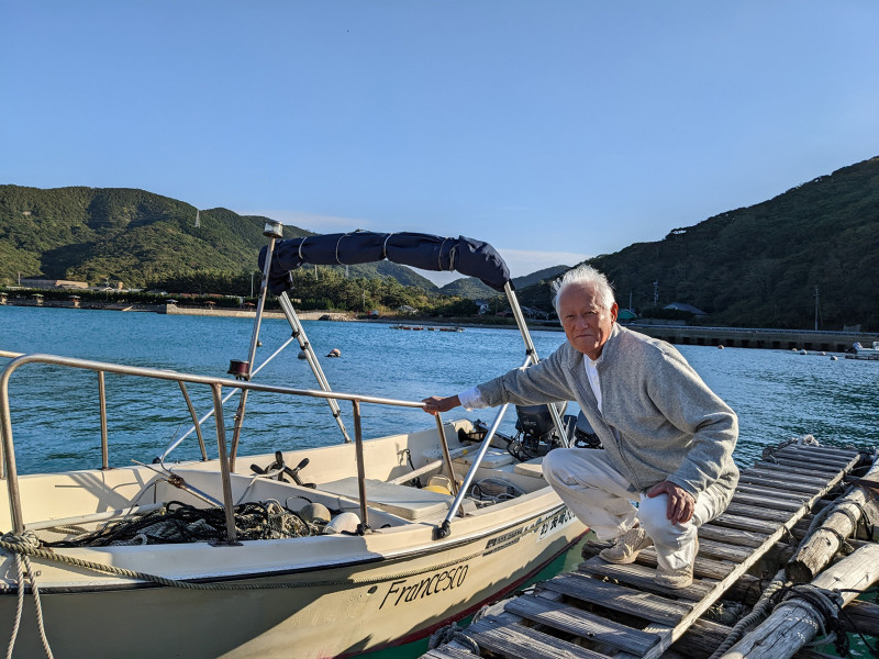 A photograph of Kakimori Kazutoshi with boat on Naru Island. Senior Japanese man in grey jacket kneeling beside a white boat on a sunny day.