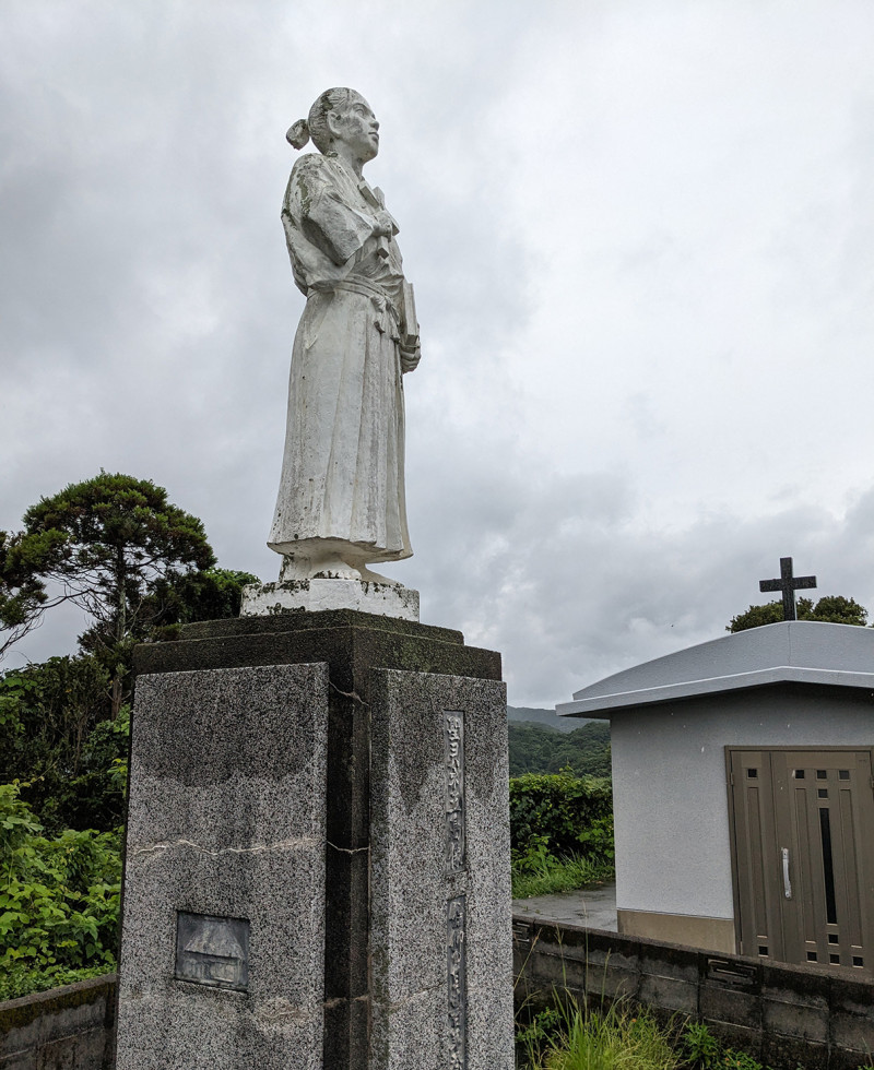 A photograph of a stone statue of Yohane Gotō found on Fukue Island at Mizu-noura Church. The statue is of a man in kimono holding a cross to his chest and a book at his side, staring into the distance.