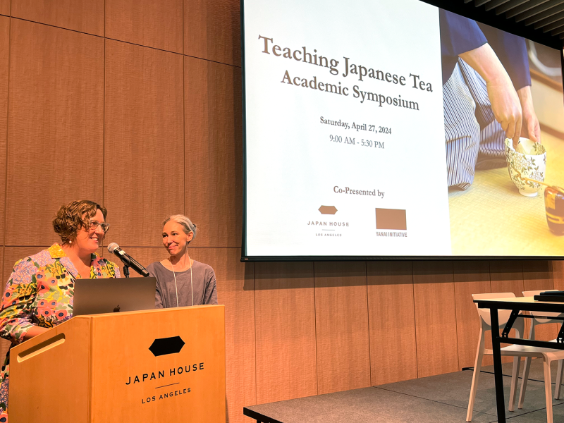 Two women stand at a podium with the label "Japan House" on it, introducing the conference for which there is a large projection that says "Teaching Japanese Tea Academic Symposium."