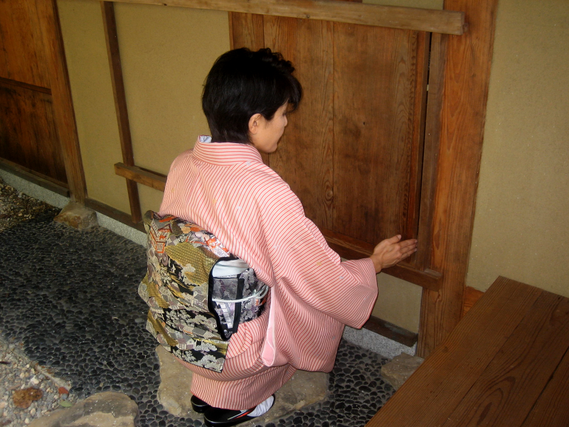 A Japanese woman in a pink and white vertical striped kimono and gold and black patterned obi kneels in front of the door to a tea house. Her back is to the viewer and her hand on the door of the tea house, about to open it.