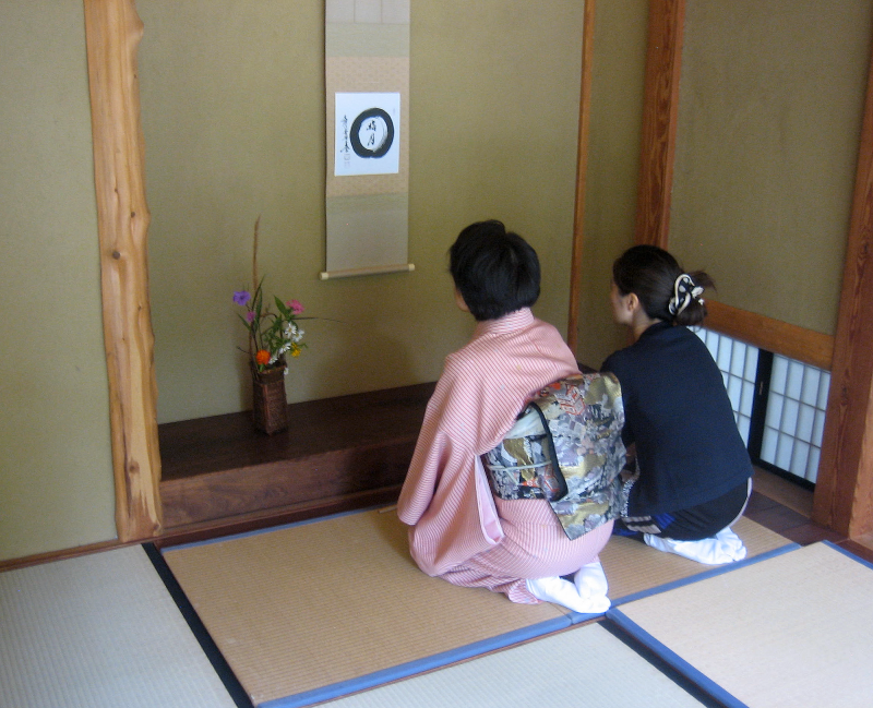 Two Japanese women kneel on tatami in front of the tokonoma, with is adorned with a hanging scroll and a flower arrangement. They are looking up at the scroll and observing the circular painting with calligraphic writing on it.