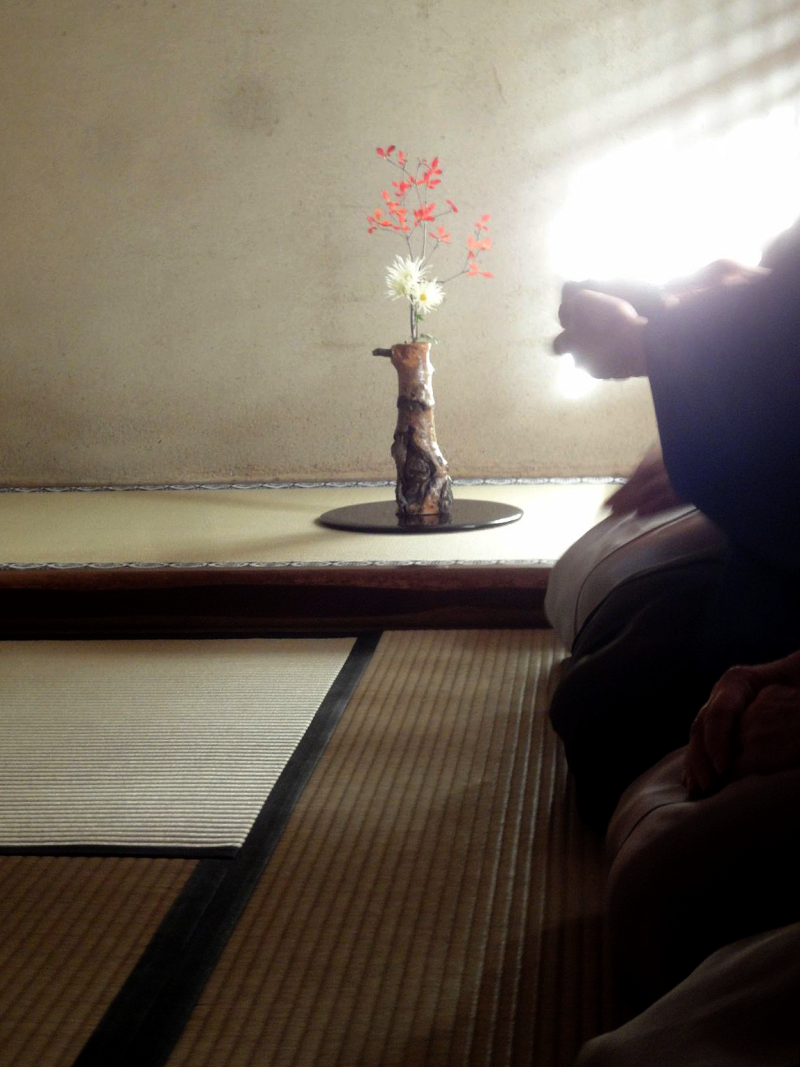 A shadowy photograph of tea practitioners sitting before a tokonoma with flowers set up in it. They are cloaked in shadow and only their knees are visible on the right-hand side of the photo, with the person farther away holding up a bowl of tea.