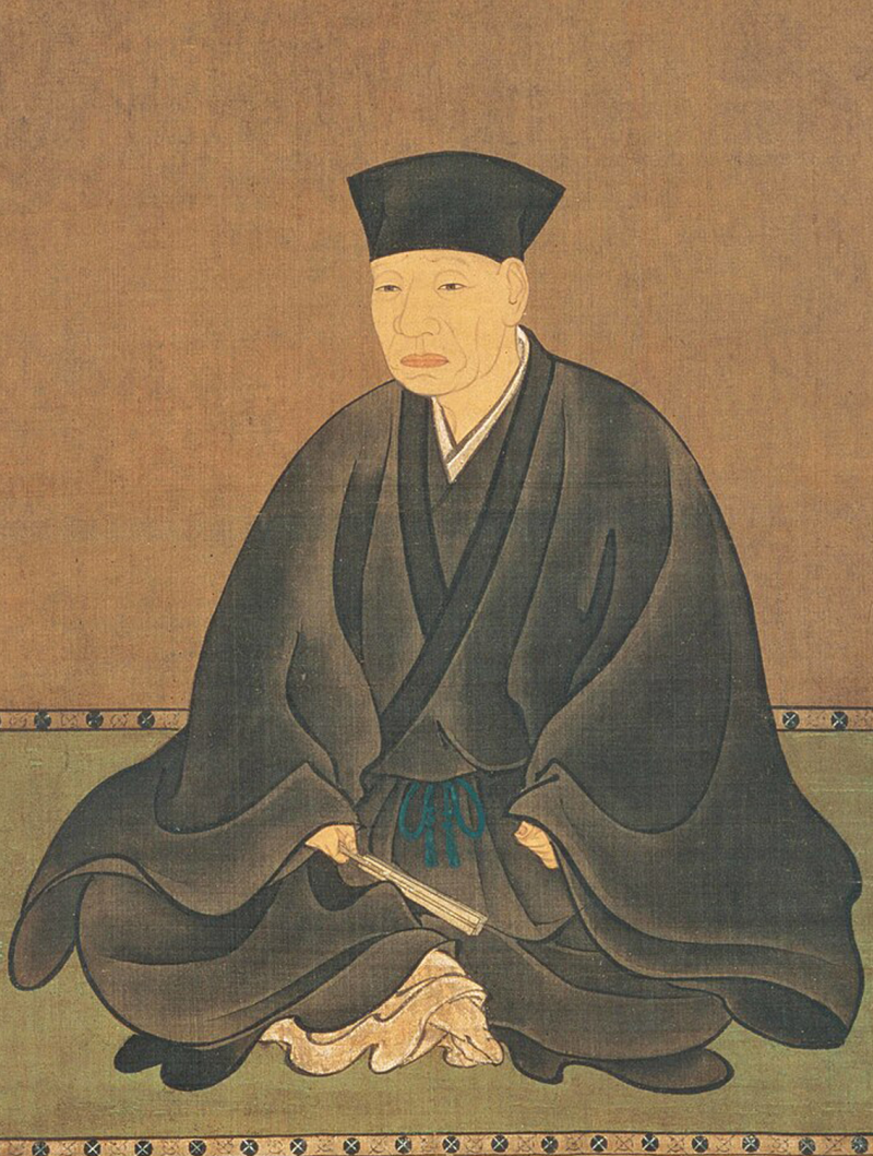 A man with a drawn face sitting in formal black Japanese clothes with a black cap on, holding a fan in one hand.