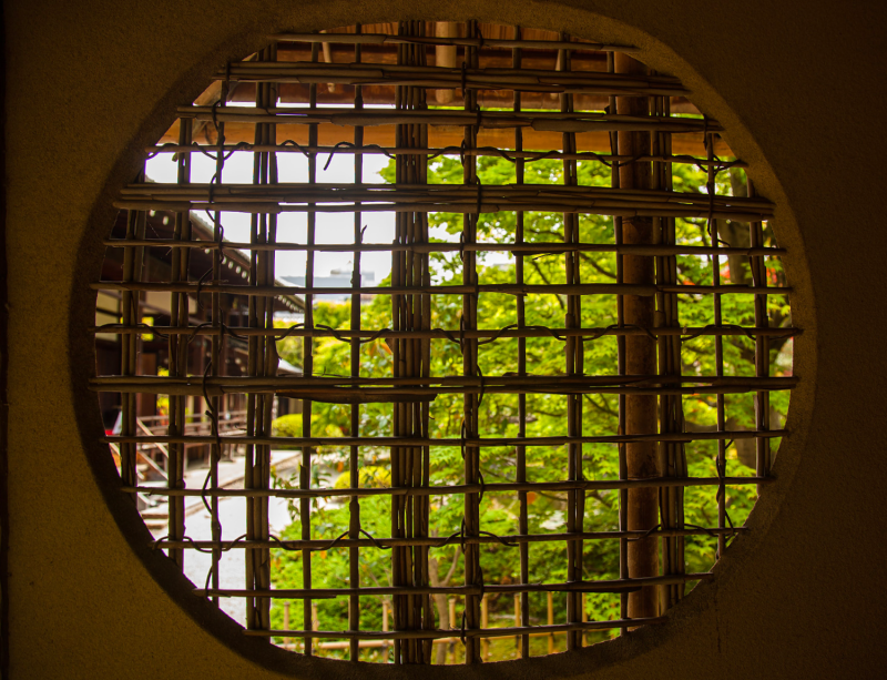 A large circulate window with latticework made of wooden stripes and vines. In the background is greenery and a wooden building.