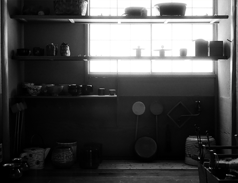 A black and white photo of a preparation room with a bright window in the background and several shelves with various tea utensils spread throughout.