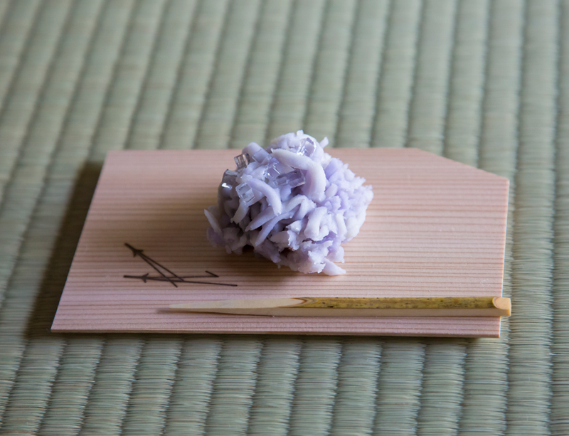 A small lavender-colored tea sweet that looks like a many-petaled flower blossom, resting on a small piece of wood with a bamboo pick next to it.