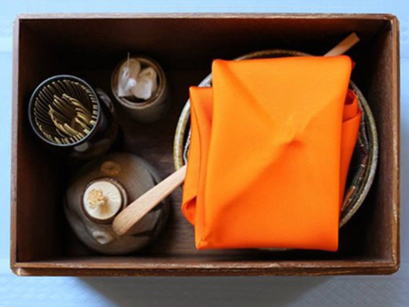 An open wooden box with a small tea container containing matcha placed inside a teabowl, above which is a diagonally placed tea scoop and then a folded silk wiping cloth on top. At the top left of the box is a ceramic jar containing small sweets, next to it, on the right, are cylinders holding a bamboo whisk and a damp linen cloth.