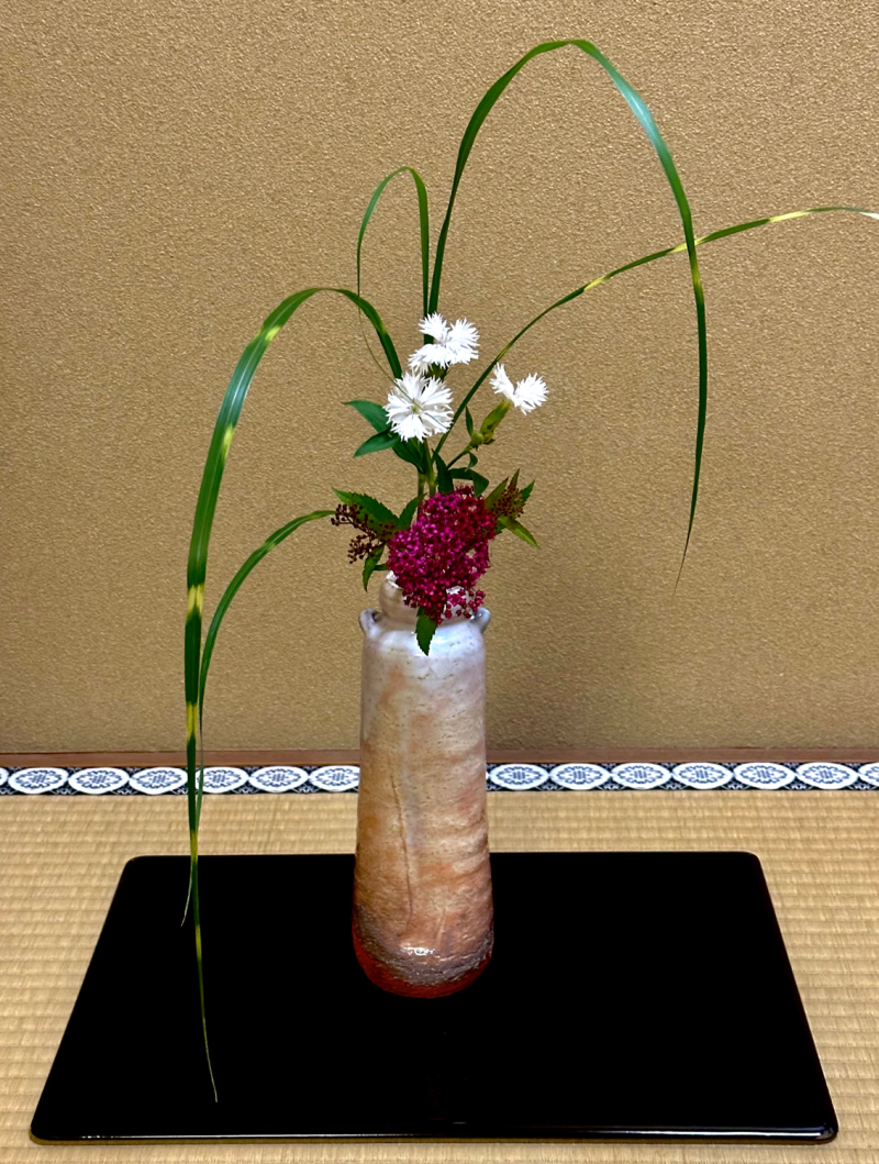 A brown ceramic vase placed on top of a black lacquer board holds white and pink flowers with green leaves.