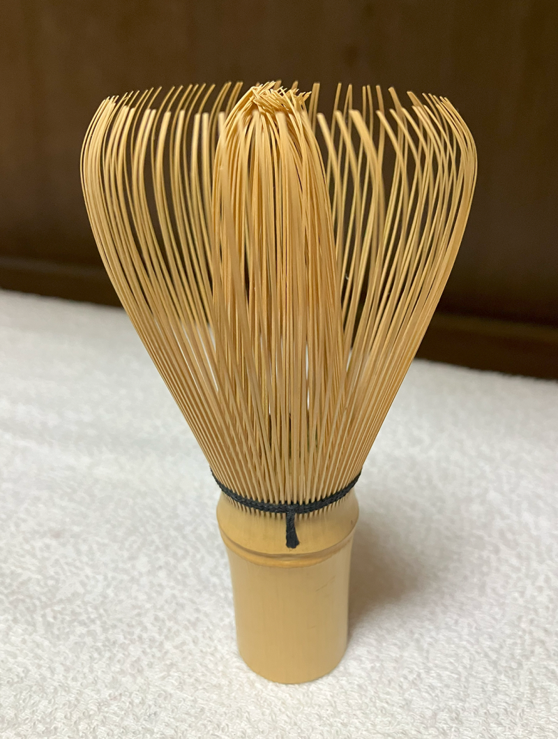 A bamboo tea whisk, standing on its back with the thin whisk tendrils pointing upwards.