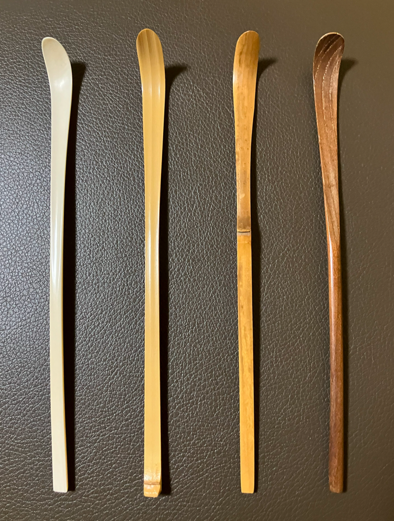 Four long thin spoons for matcha, arranged on a pebbled surface. From left to right, an ivory (or imitation ivory) carved scoop, pale tan bamboo with node at the base of the handle, the most common pale tan bamboo with node centered in the handle, and a brown wooden carved scoop with visible woodgrain.