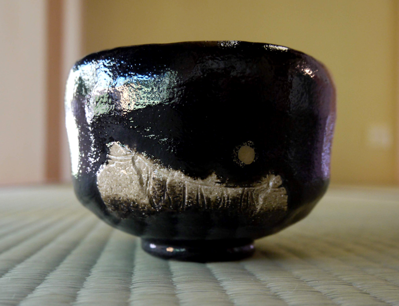 Black, shiny Raku style tea bowl with an irregular pale front marking, sitting on tatami reed mat.