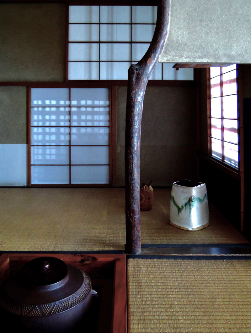 The corner of a traditional tea room, featuring a shortened tatami mat before a sunken hearth with tea utensils resting on the mat. There are small shoji-covered windows in the background and a short divider made of wood next to the hearth.
