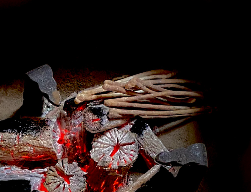 an interior view of a brazier. On the top right of a carefully arranged pile of black charcoal are a bundle of white twig charcoal sticks. The fire is burning and all the charcoal is burning, the white twigs have begun to to take on a burnt hue and the black charcoal are turning white.