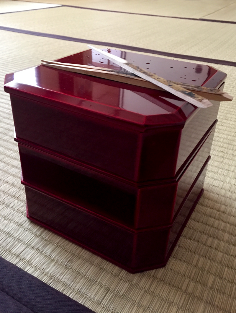 Three vermillion colored square lacquer boxes srae stacked on top of each other and sitting on a tatami floor. The top of the stack of boxes has a lid on which are laying three wood picks, used for picking up the sweets housed inside the boxes. There are two sticks laying horizontal and a third is placed diagonally, resting over the other two sticks. A light sprinkling of water creates a shimmer on the lacquer lid.