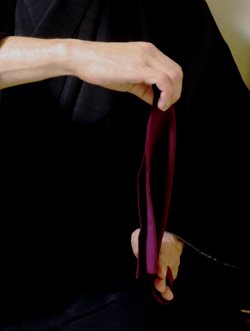 A man wearing black Japanese clothes is holding a purple square silk cloth in front of him with both hands, it is held vertically and he is in the process of folding it in half into a triangle shape.