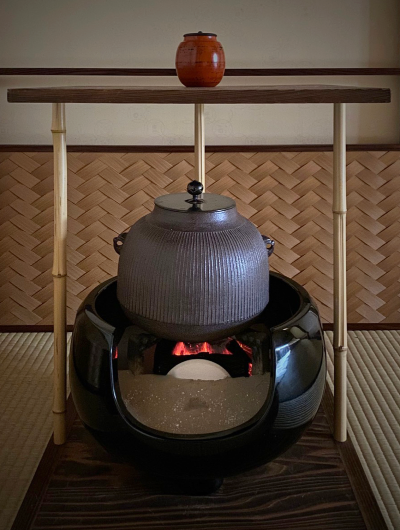In the background there is a woven screen, in front stands a cast iron kettle resting on a cast iron stand set into a bed of ash and charcoal that fills a black lacquer brazier. The brazier is round in shape with a low cut out "mouth" in front allowing a view of the fire. The brazier itself sits on the bottom shelf of a two shelved wooden utensil stand. The top shelf holds a deep red tea container.