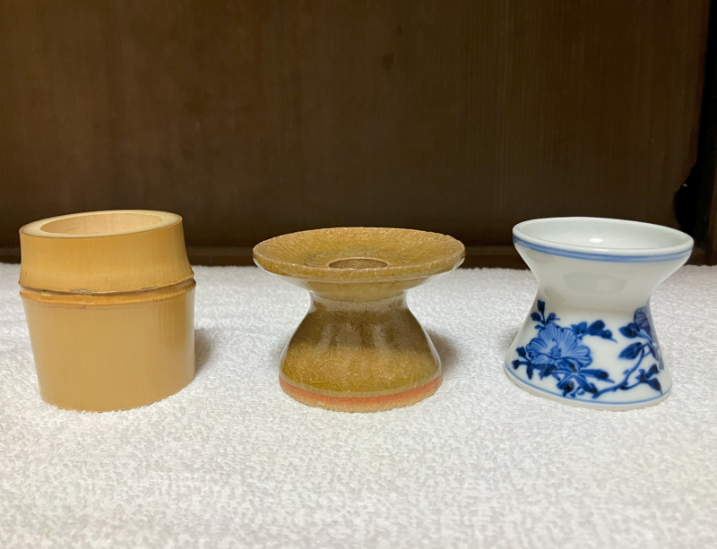 Three examples of lid rests. On the left, a pale aged bamboo, cut with one node in the top half of the item. In the middle, a yellow ceramic glazed piece that narrows in the center and widens to a disc-shaped top. On the right, blue and white picture porcelain with a gentler narrowing in the center.