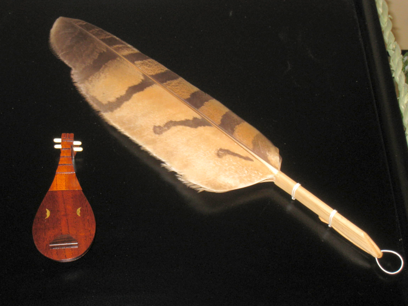A large tan-colored feather with brown stripes and a wooden handle resting on a black tray next to an incense burner shaped like a tiny biwa.