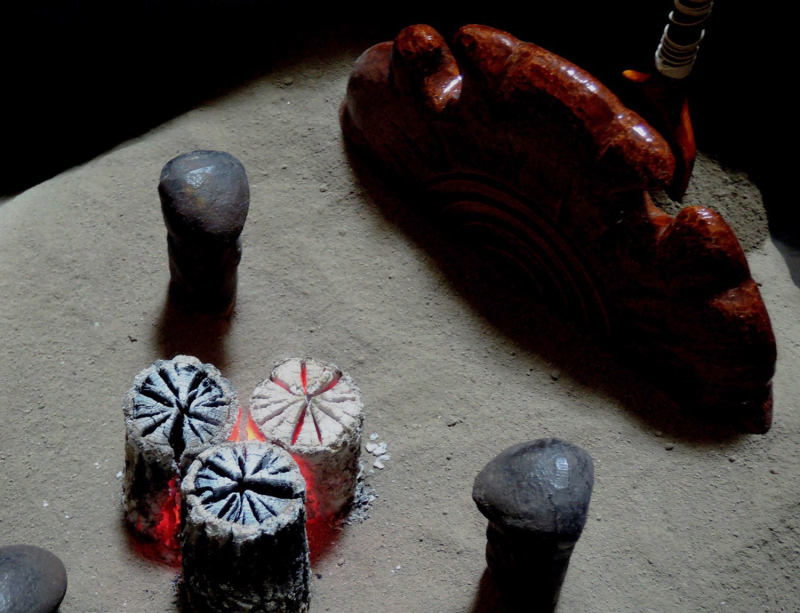 An interior view of the braizer shows a bed of grey ash into which are placed three sticks of black charcoal, with a faint red glow and the tops hjave started to turn white, and a half circle brown disc in the far back of the braizer is sunk into the ash. The three feet of a cast iron trivet to hokd the kettle are visible just above the bed of ash, they form a ring around the charcoal.