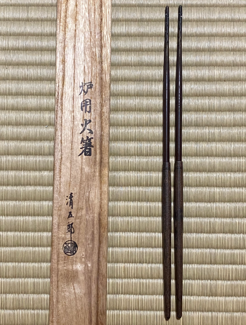 On the right are two long metal chopsticks, on the left is a wooden storage box for them.