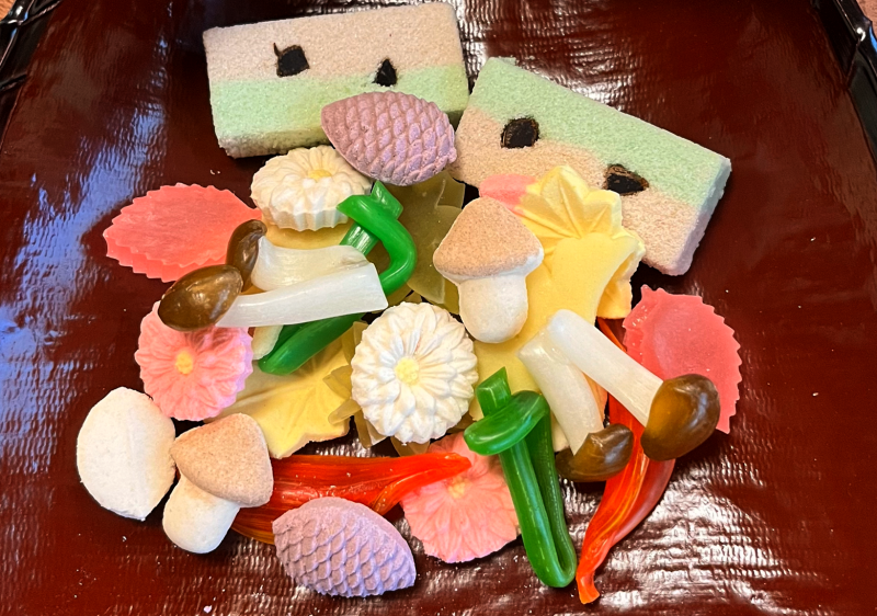 A pile of various, multicolored dry sweets shaped like flowers, mushrooms, pine cones, and other small objects, resting in a lacquered dish.